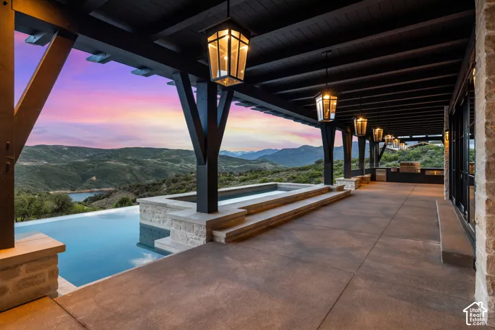 Expansive covered patio with a built-in outdoor kitchen, infinity-edge pool, and integrated spa, all set against a backdrop of distant mountains and a lake