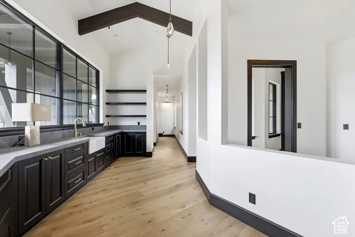 Vaulted ceiling with exposed wood beam, wood-finish flooring, built-in cabinetry with stone countertops, farmhouse sink, and large grid-pane windows