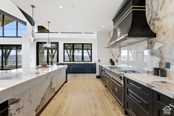 Spacious kitchen featuring an expansive island with a waterfall edge, light wood-finish flooring, dark cabinetry, and a professional-grade range with a custom hood