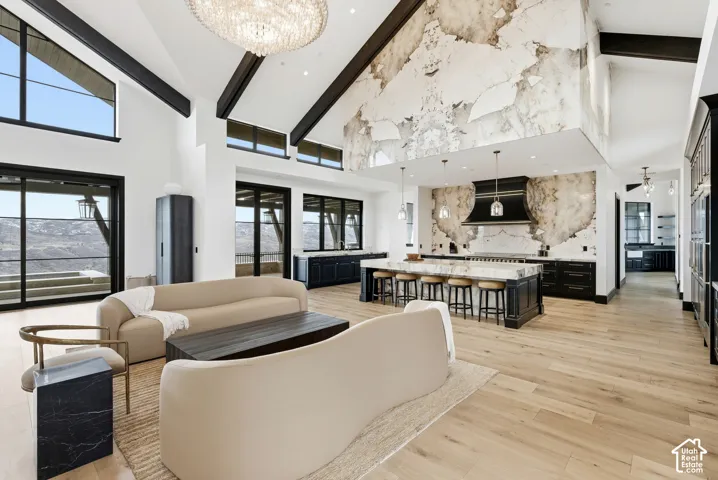 Vaulted ceilings with exposed beams, expansive windows, light wood-finish flooring, a large kitchen island with seating, and a distinctive textured accent wall