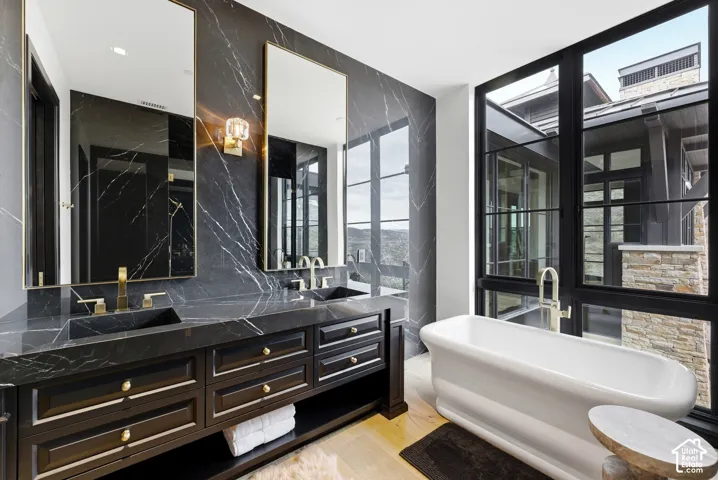 Freestanding soaking tub, black framed windows, dual vanity with undermount sinks, stone slab accent wall, and wood-finish flooring