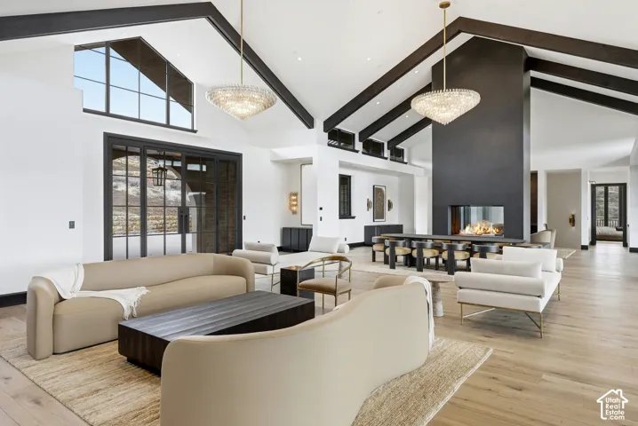 Vaulted ceiling with exposed beams, wood-finish flooring, two large crystal chandeliers, a built-in fireplace, and expansive windows