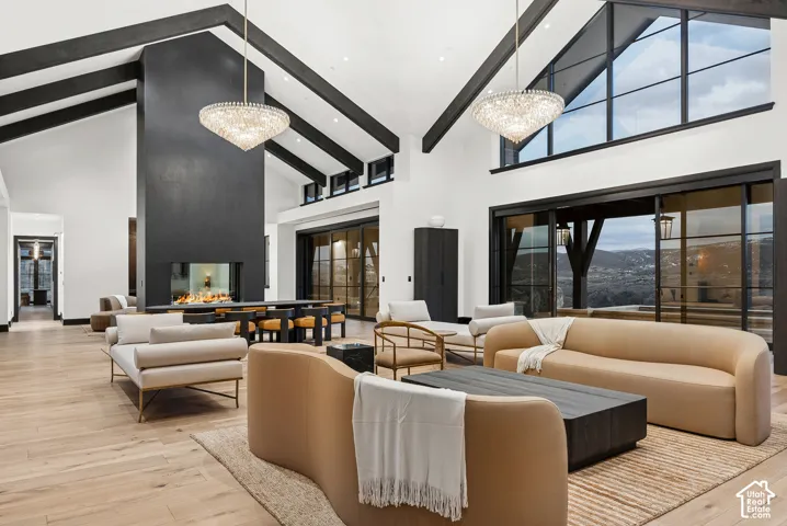 Vaulted ceiling with exposed dark beams, expansive black-framed windows, large central fireplace feature, wood-finish flooring, and dual crystal chandeliers