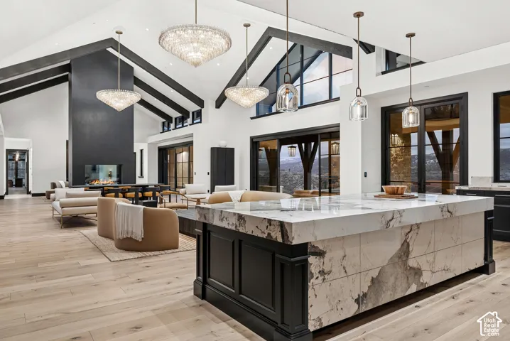 Vaulted ceilings with exposed beams, central fireplace feature, expansive kitchen island with a waterfall countertop, wood-finish flooring, and multiple tiered chandeliers