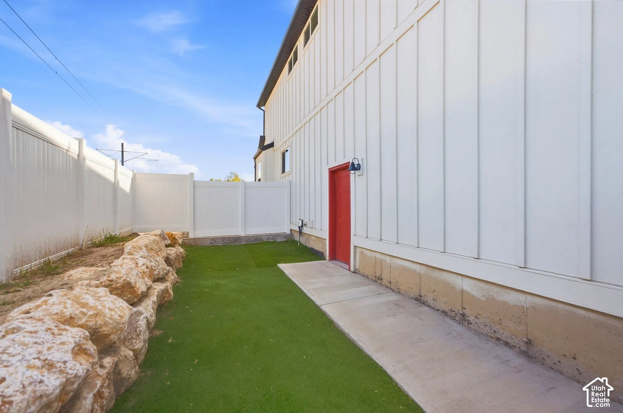 Private outdoor space. White vertical siding exterior with a red entry door, artificial turf, concrete pathway, and a natural stone retaining wall