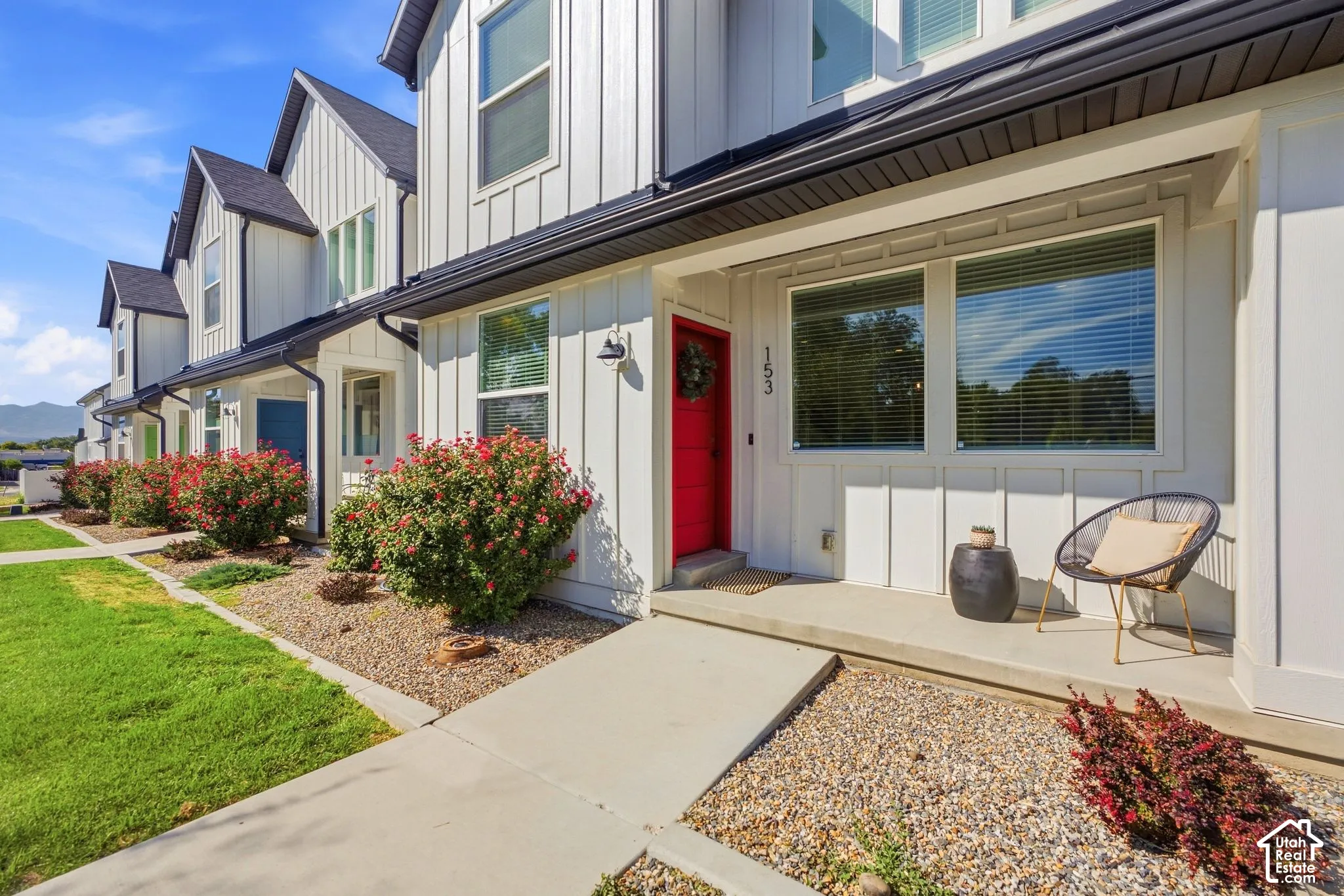 White vertical siding exterior with black trim and gutters, featuring a red entry door, covered front porch, and front landscaping with shrubs and gravel