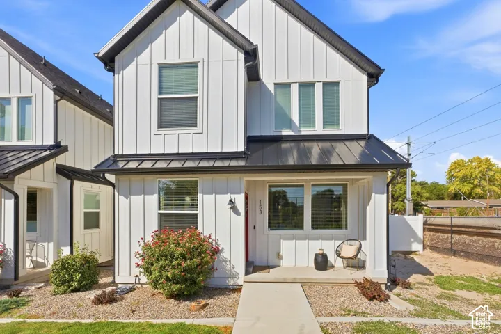 Board and batten siding, black metal roof accents, covered entry, concrete pathway, and established plantings