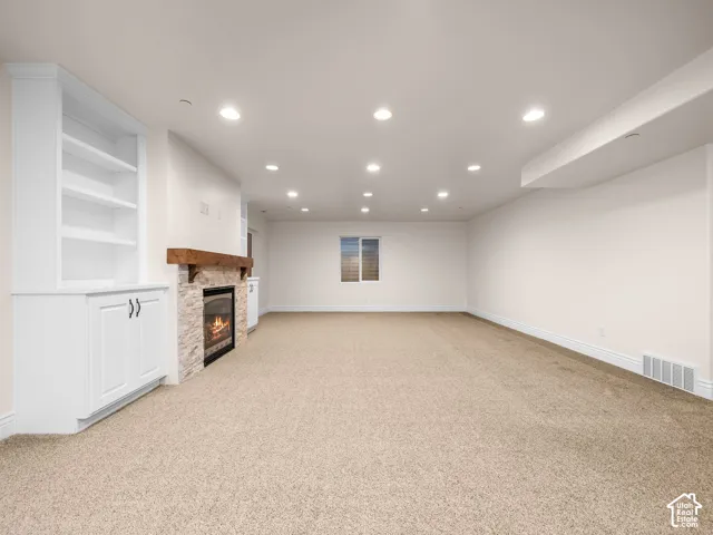 Spacious room featuring carpet flooring, built-in shelving, a stacked stone fireplace with a wood mantle, recessed lighting, and a window