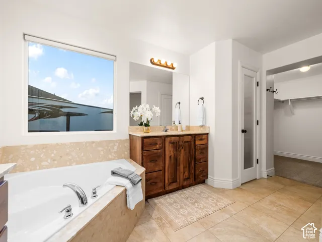 Built-in soaking tub with polished chrome fixtures, dual vanity with wood cabinetry and stone countertop, large window, and tiled flooring