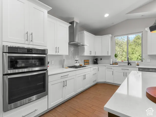 White shaker cabinetry, dual stainless steel ovens, wood-finish flooring, white countertops, and a stainless steel range hood