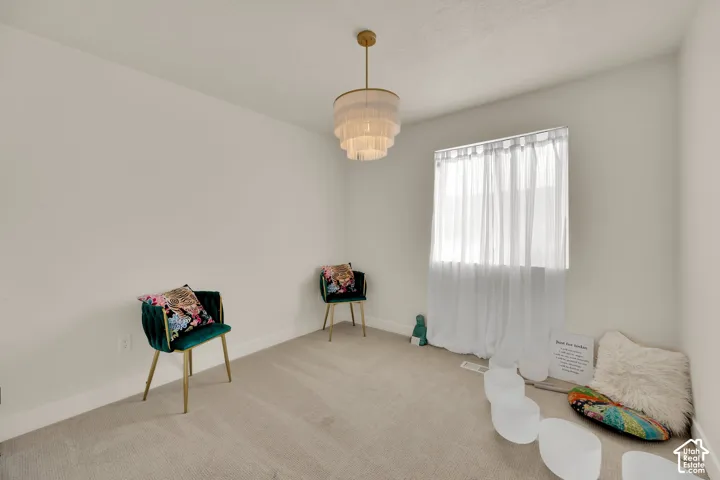 Neutral wall paint, carpeted flooring, a tiered chandelier, and a window with sheer curtains