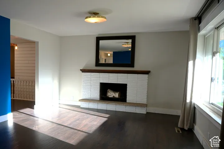 Hardwood-style flooring, white brick fireplace with dark wood mantel, window with white trim, flush-mount ceiling fixture, and a partial view of a shiplap wall