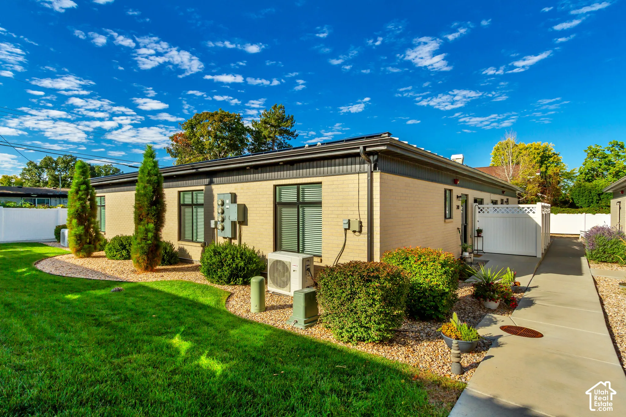 Brick exterior with black-framed windows, a concrete walkway, a white privacy fence, and manicured landscaping featuring mature shrubs