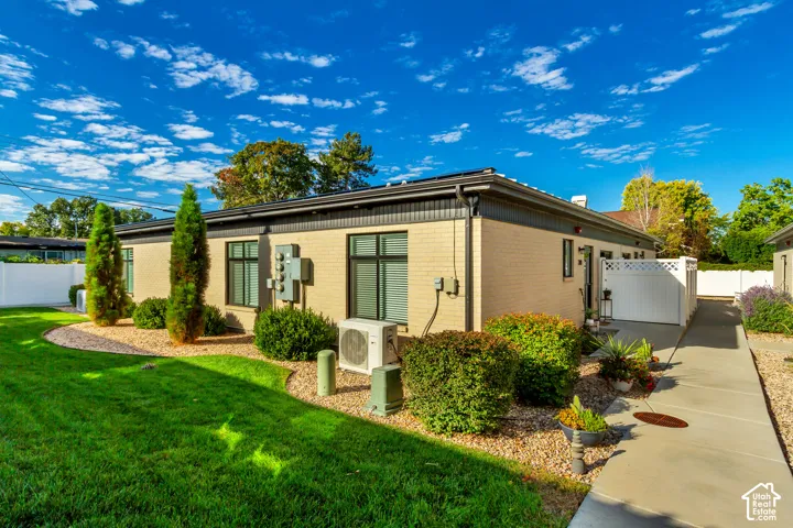 Brick exterior with black-framed windows, a concrete walkway, a white privacy fence, and manicured landscaping featuring mature shrubs