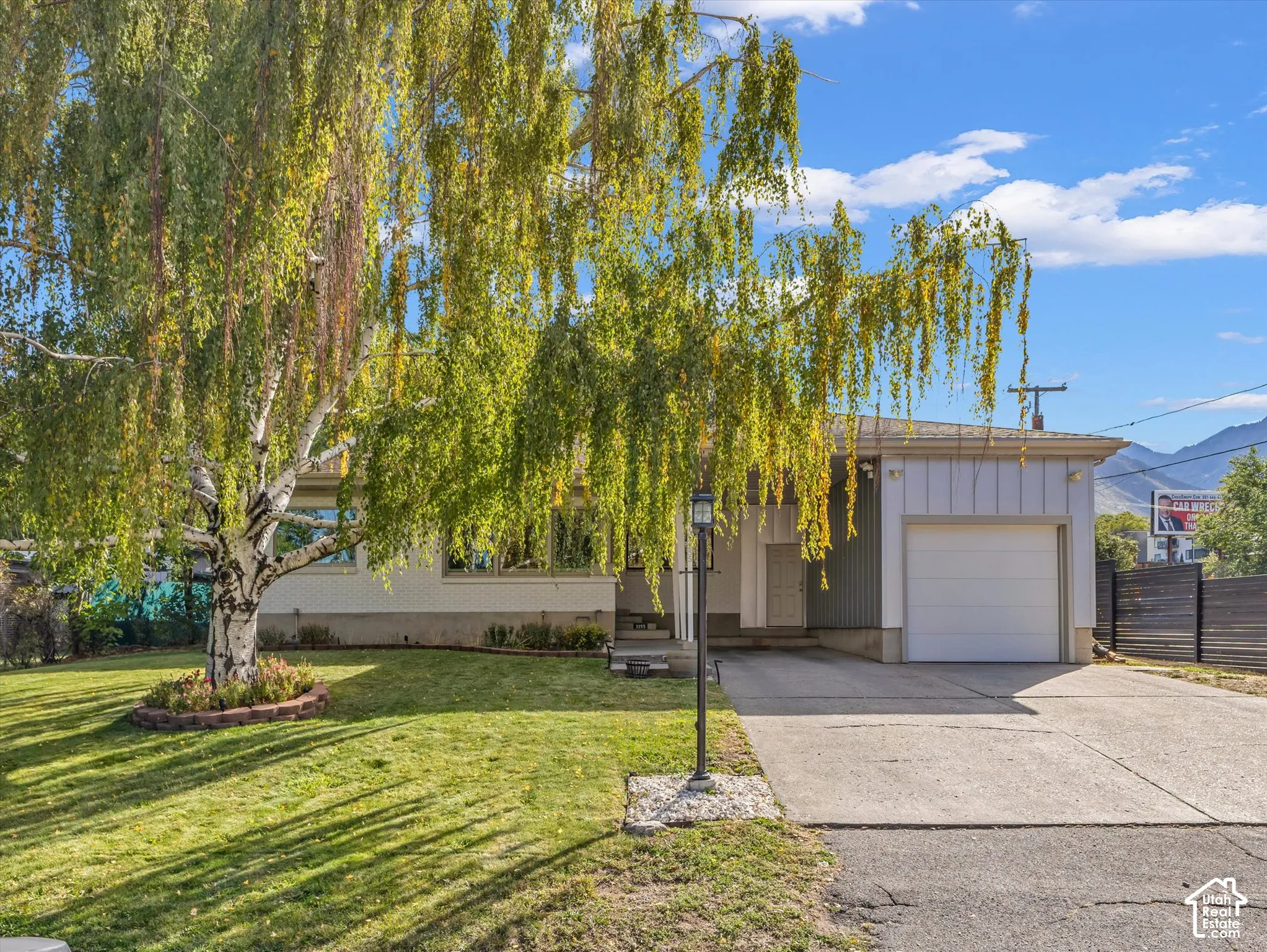 Single-story exterior with a side-facing garage, concrete driveway, and a front lawn featuring a mature tree with a brick-lined bed