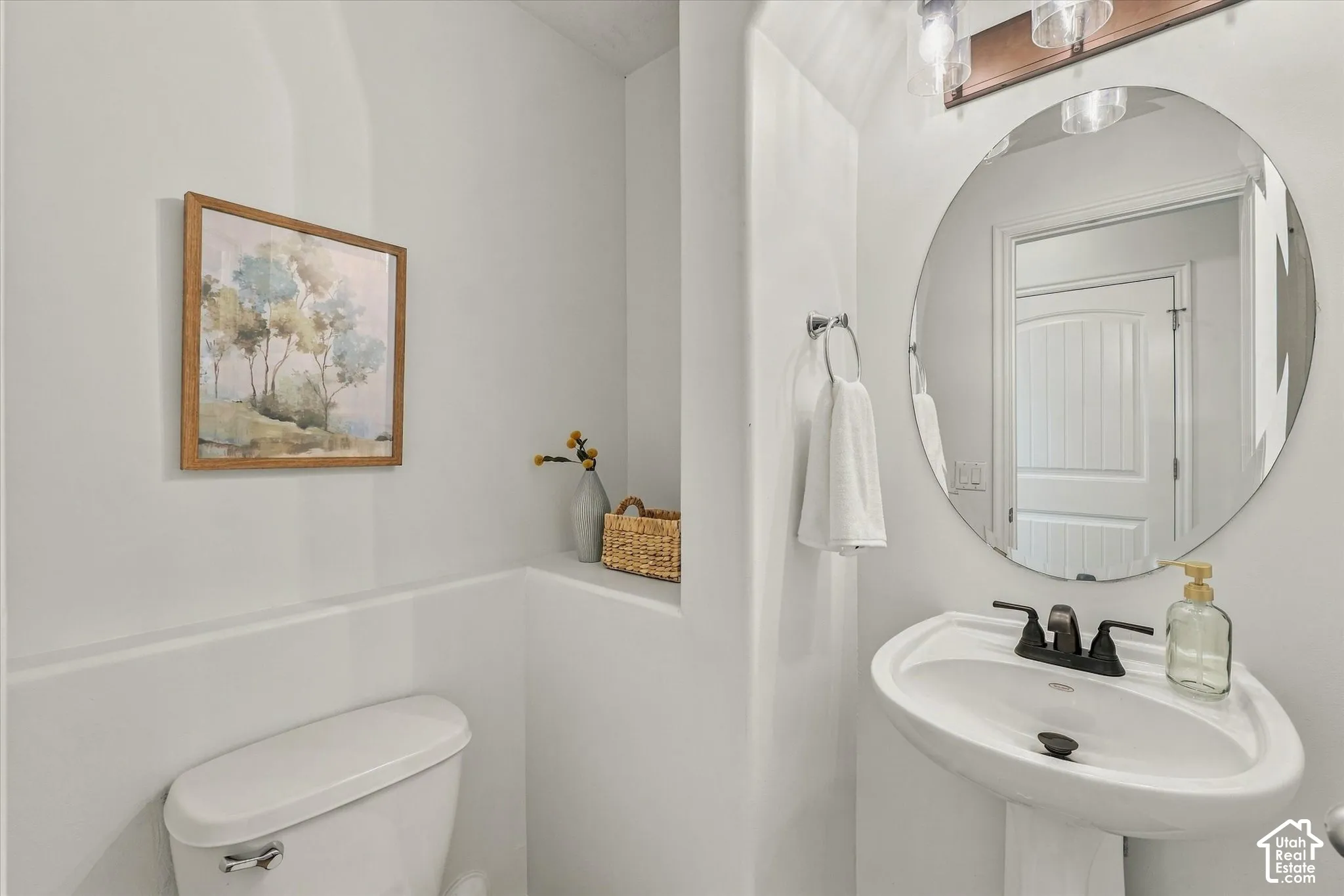 Pedestal sink with a matte black faucet, wall-mounted circular mirror, and overhead vanity lighting
