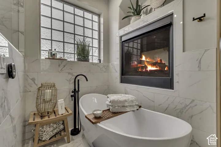 Freestanding soaking tub with a black floor-mounted faucet, complemented by a built-in fireplace, glass block window, and marble-patterned tile surround