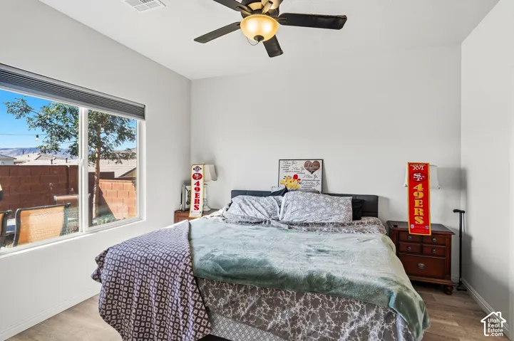 Light gray walls, wood-finish flooring, ceiling fan with integrated lighting, and a large window with roller shades