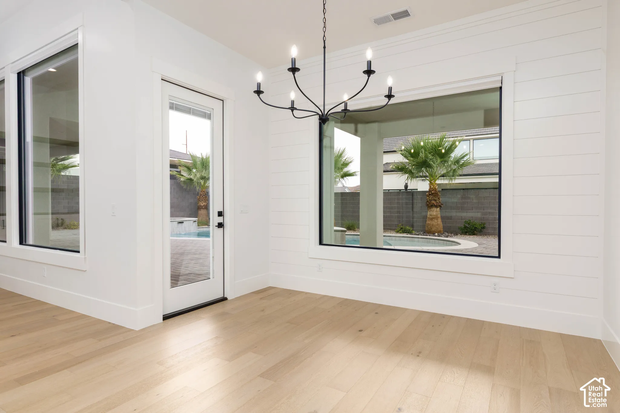 Light wood-finish flooring, white shiplap accent wall, multiple large windows, glass-paneled door, and a black metal chandelier