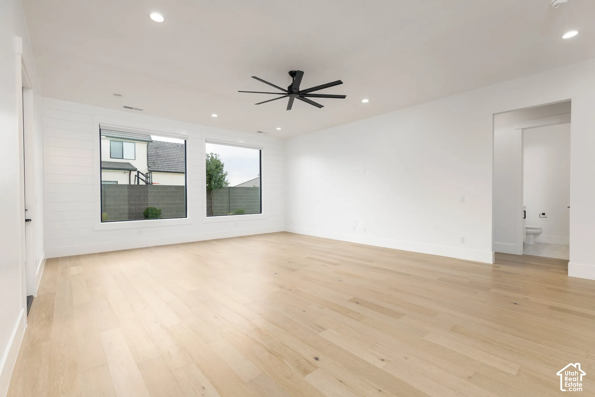 Spacious room featuring light wood-finish flooring, two large windows, a modern ceiling fan, recessed lighting, and white shiplap accent wall