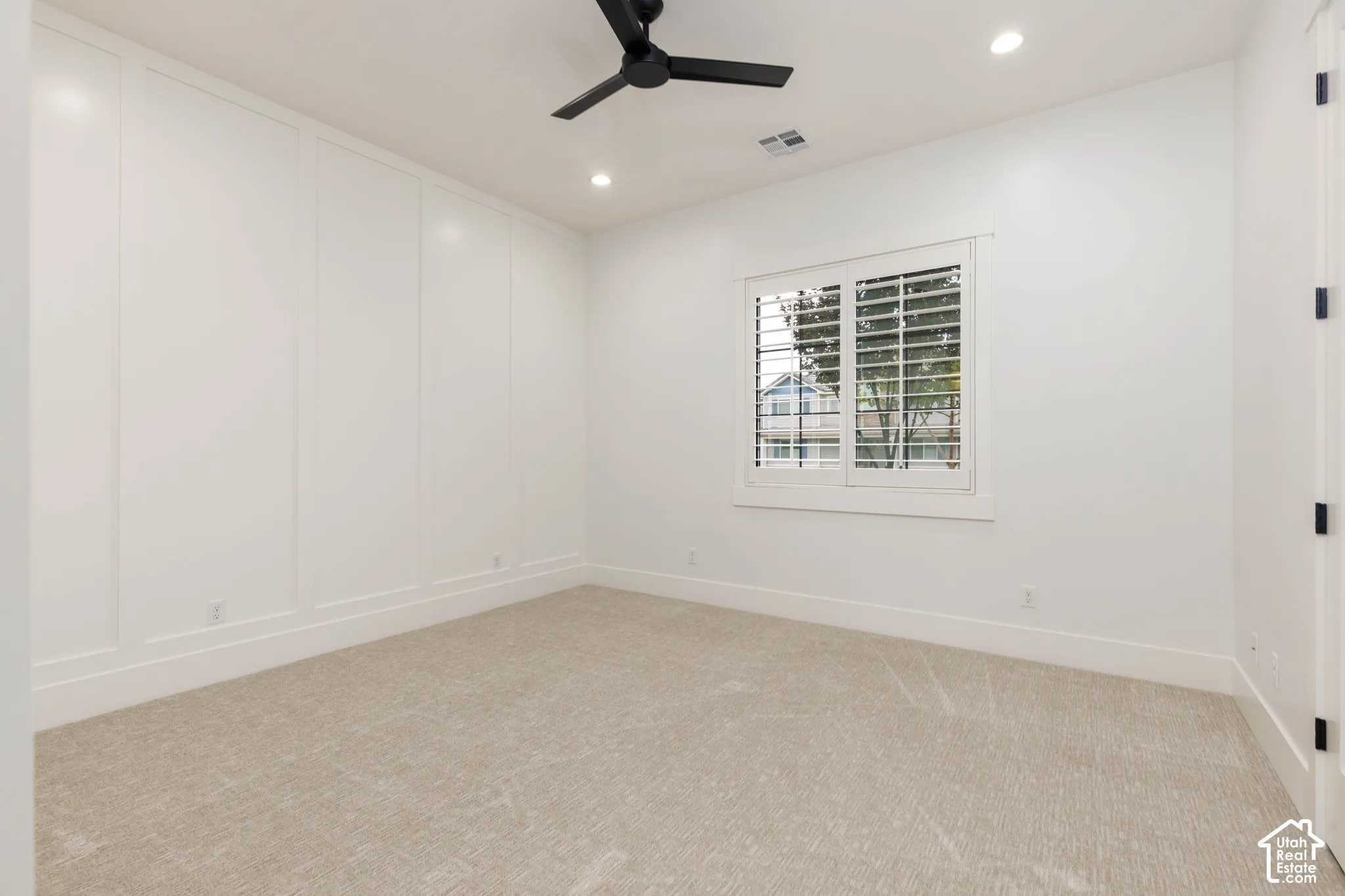 Carpeted room featuring a ceiling fan, recessed lighting, a window with plantation shutters, and an accent wall with wainscoting