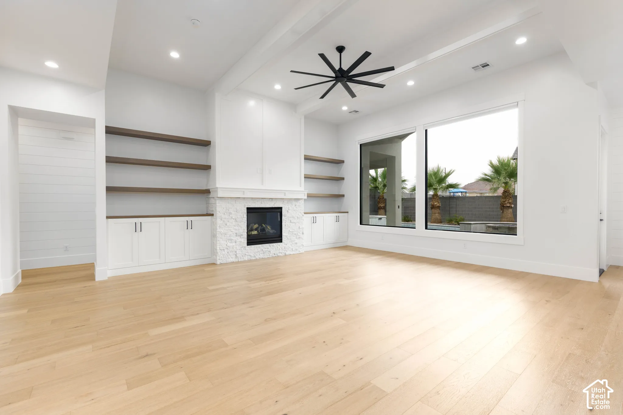 Spacious room featuring light wood-finish flooring, a stone-clad fireplace, integrated shelving with lower cabinetry, and large picture windows