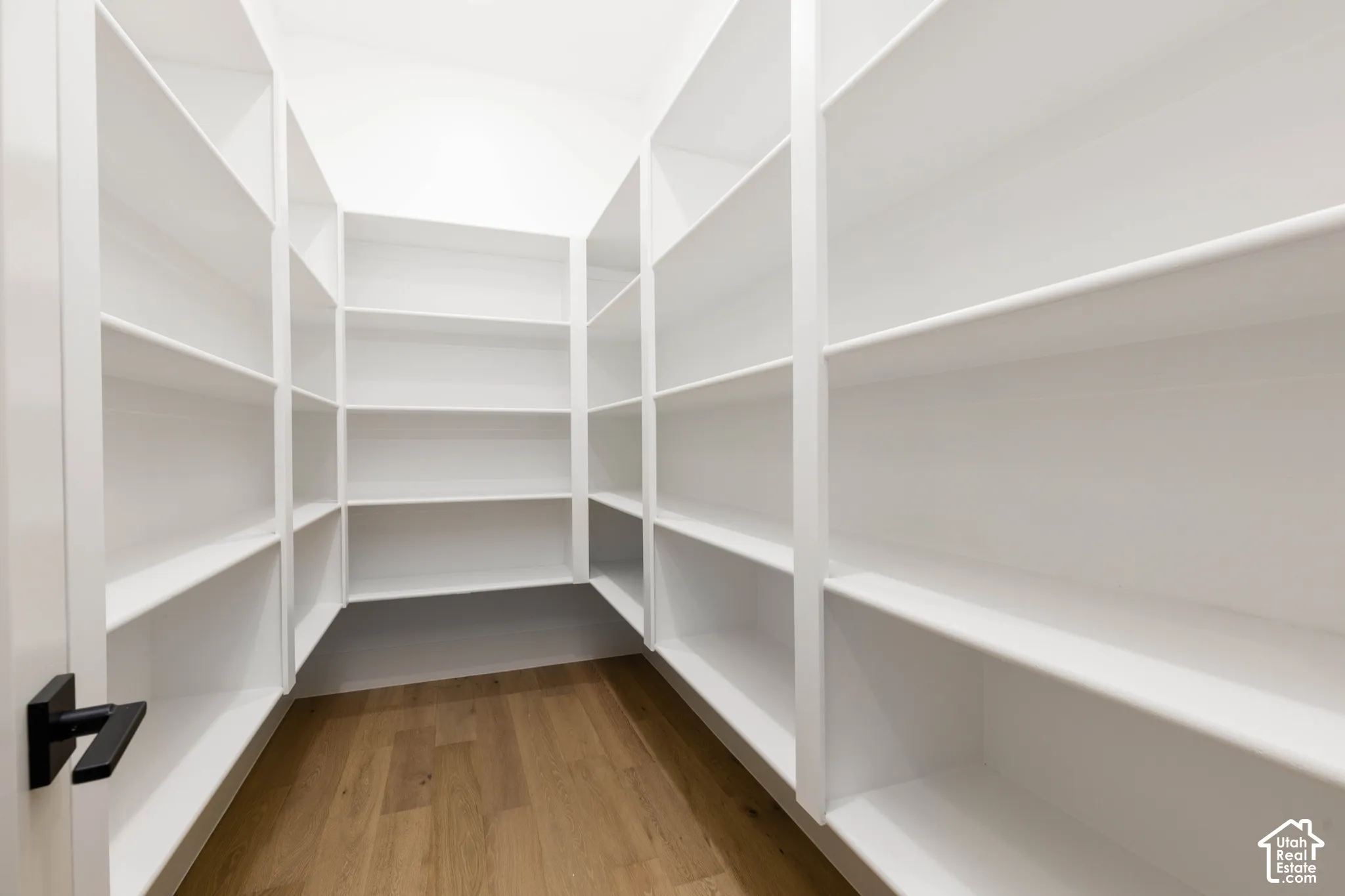 Walk-in pantry featuring extensive built-in white shelving, wood-finish flooring, and a matte black door handle