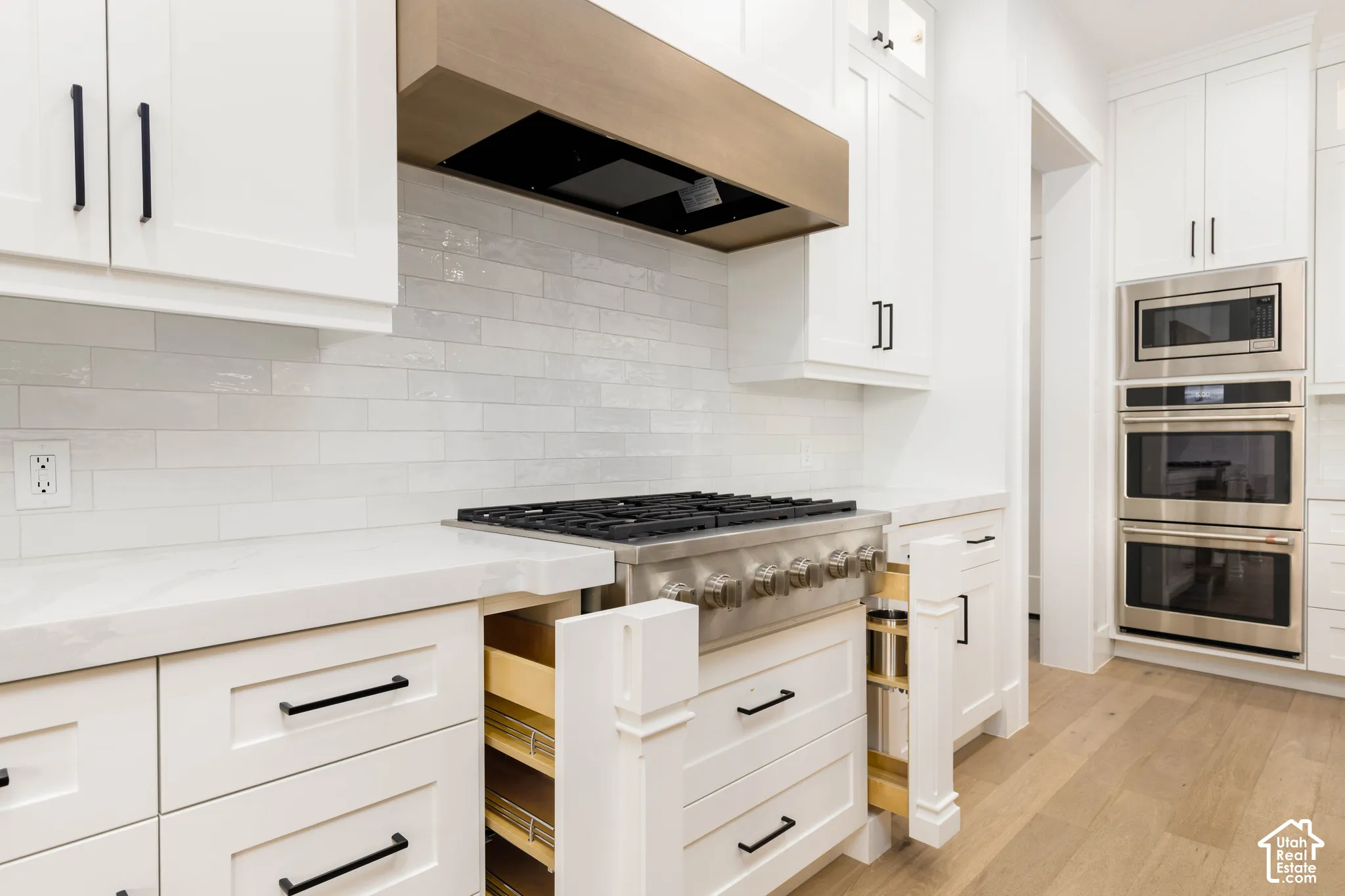 White cabinetry with black hardware, wood-finish flooring, stainless steel appliances, white subway tile backsplash, and a wood-finish range hood