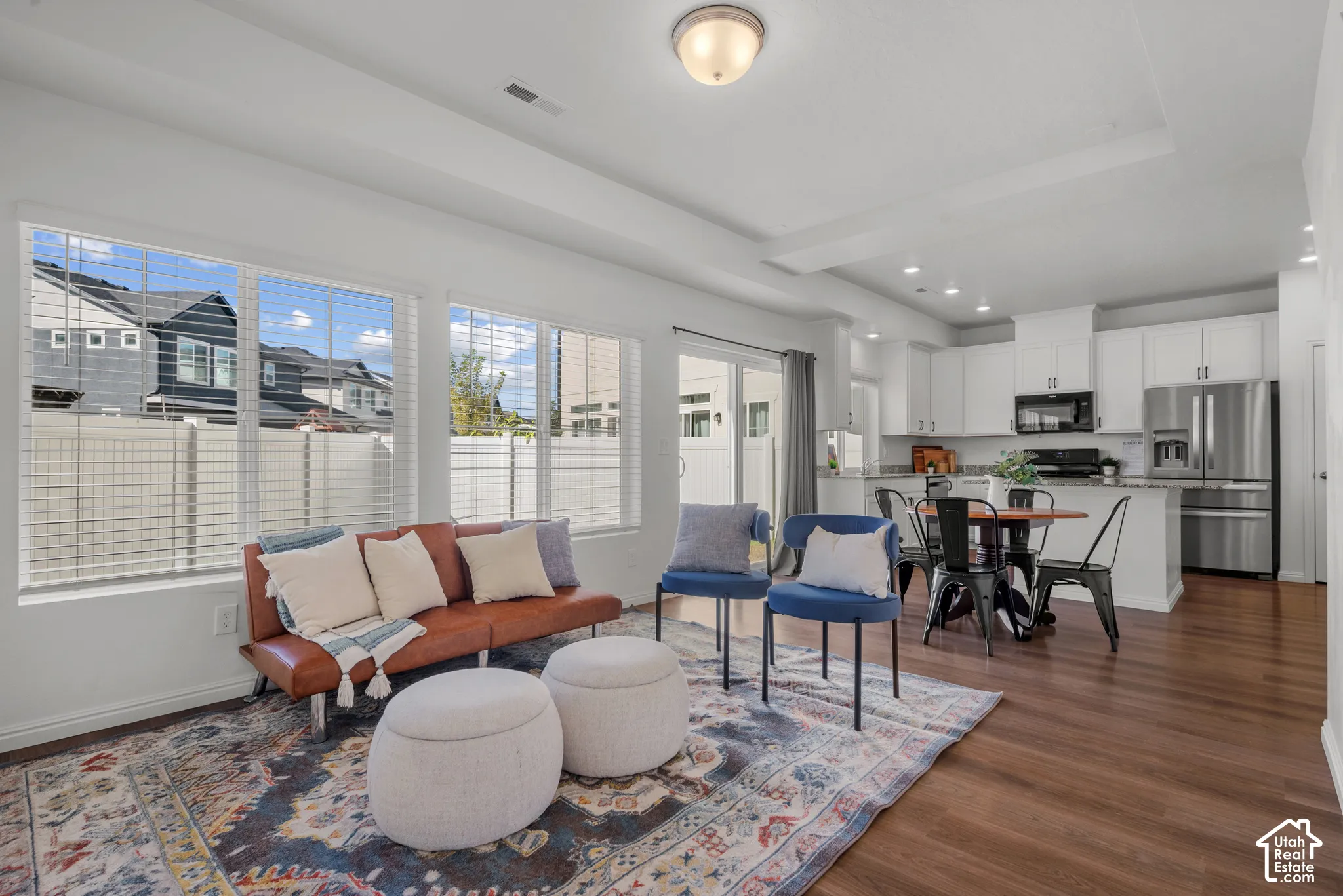 Open-concept living space featuring wood-finish flooring, white walls, large windows, and a modern kitchen with white cabinetry and stainless steel appliances, natural light,