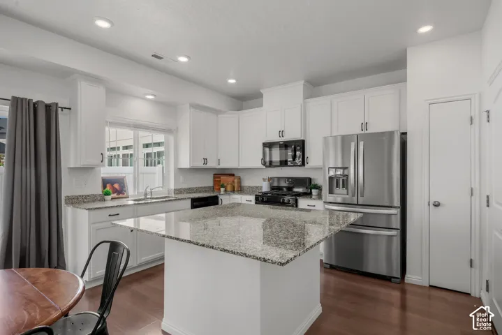 White cabinetry with black hardware, stainless steel appliances, gas stove, recessed ceiling lighting, wood-finish flooring, and a kitchen island with a granite countertop