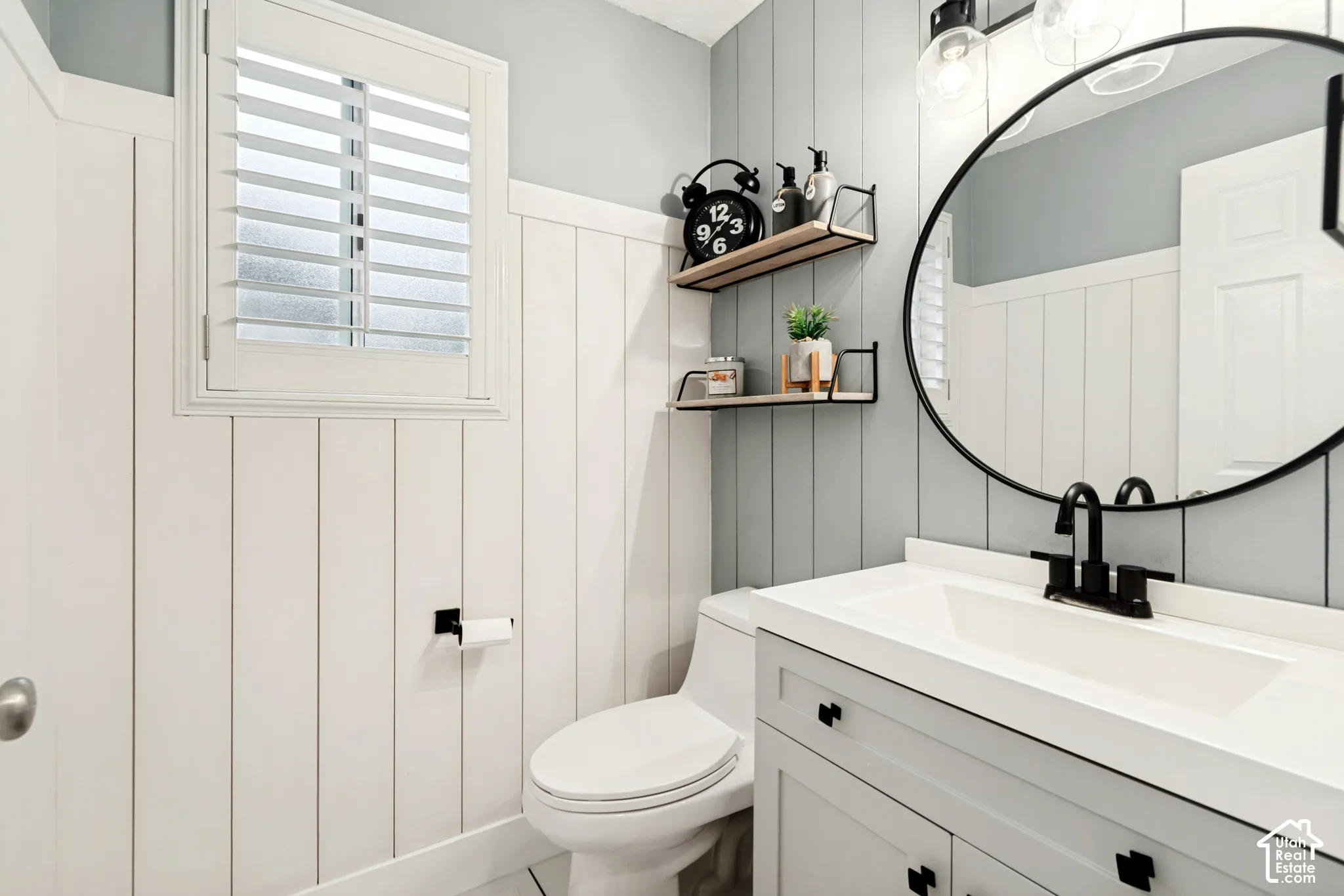 White board and batten wainscoting, white window shutters, white vanity with black hardware, black-framed round mirror, and black wall-mounted faucet