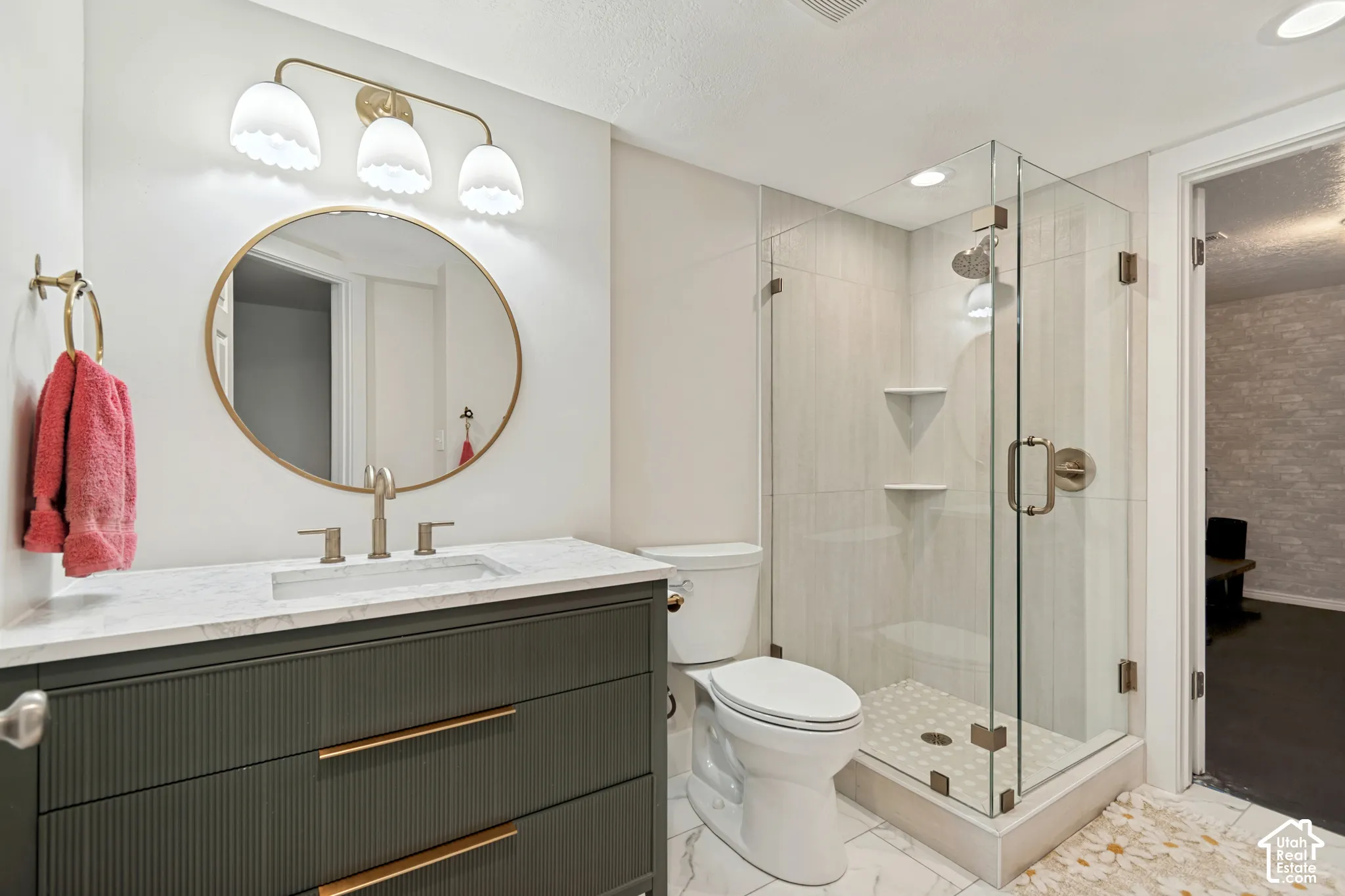 Glass-enclosed shower with a rainfall showerhead, white vanity with integrated sink and brass hardware, and a round brass-framed mirror