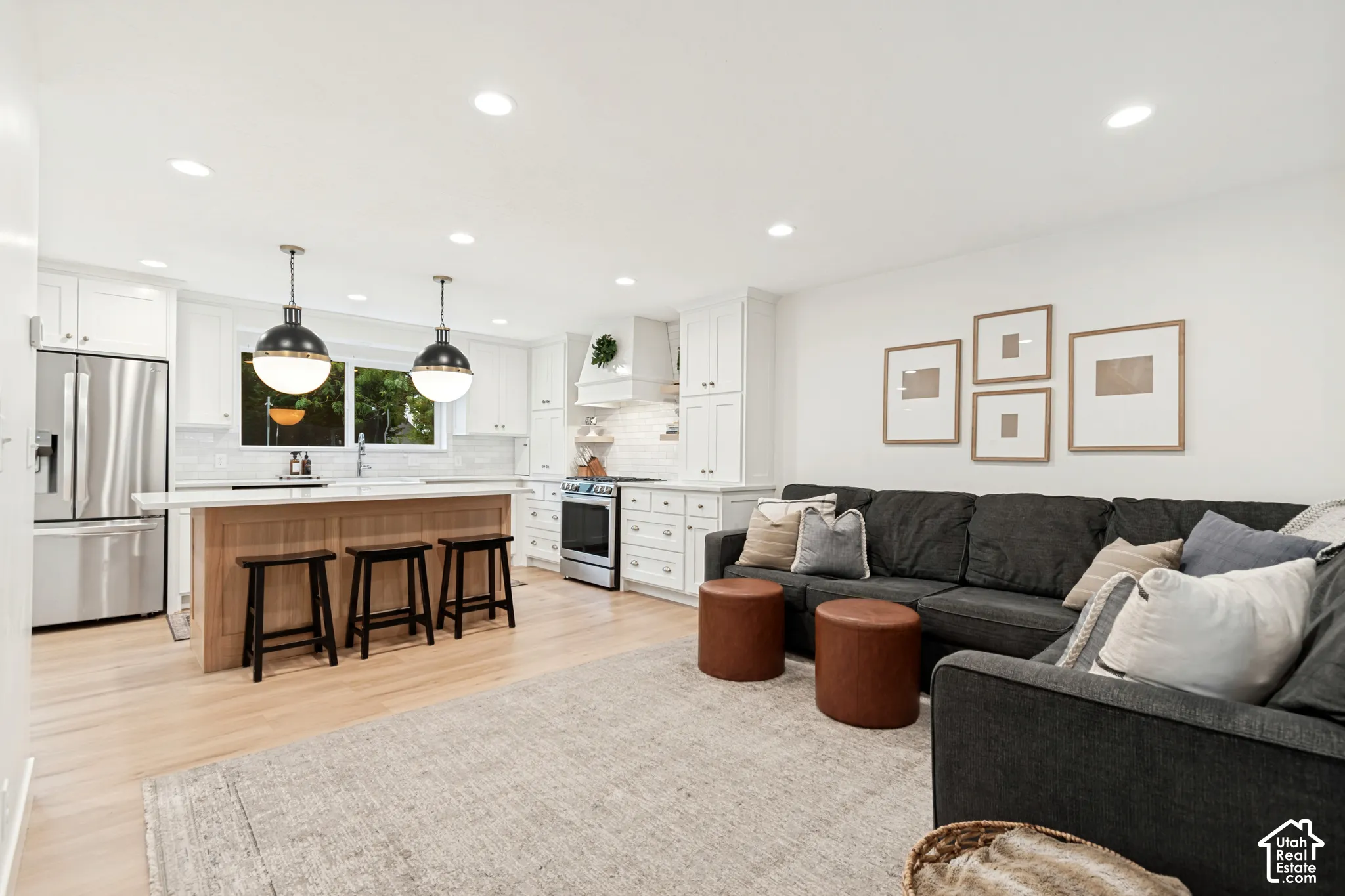 Open-concept living space featuring light wood-finish flooring, recessed ceiling lighting, a kitchen island with seating, white cabinetry, and stainless steel appliances