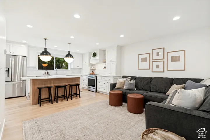 Open-concept living space featuring light wood-finish flooring, recessed ceiling lighting, a kitchen island with seating, white cabinetry, and stainless steel appliances