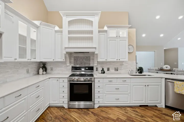White cabinetry with glass-front accents, stainless steel range and dishwasher, light-toned stone backsplash, and wood-finish flooring