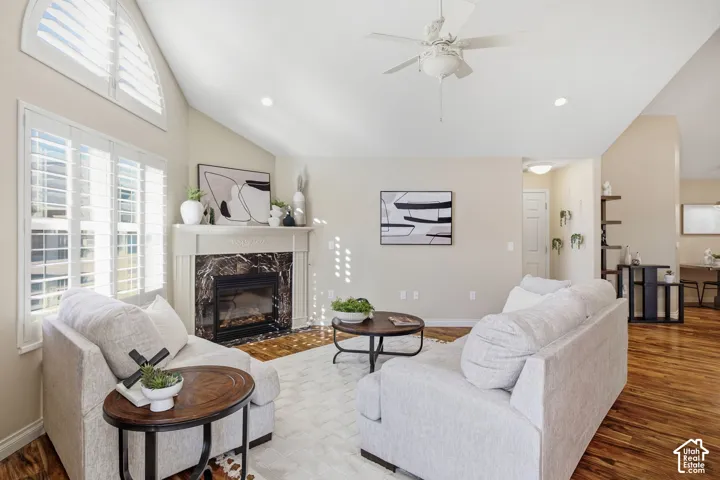 Vaulted ceiling with a ceiling fan, fireplace with a decorative mantel and dark stone surround, wood-finish flooring, and white plantation shutters