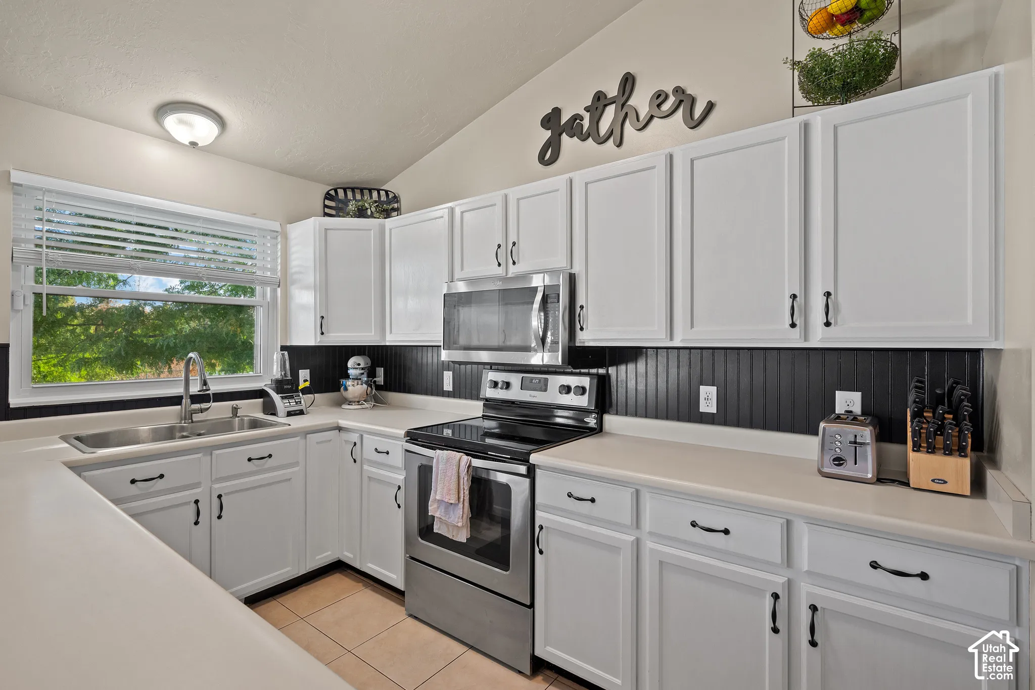 White cabinetry with black hardware, stainless steel appliances, corner sink with window, tile flooring, and a beadboard backsplash