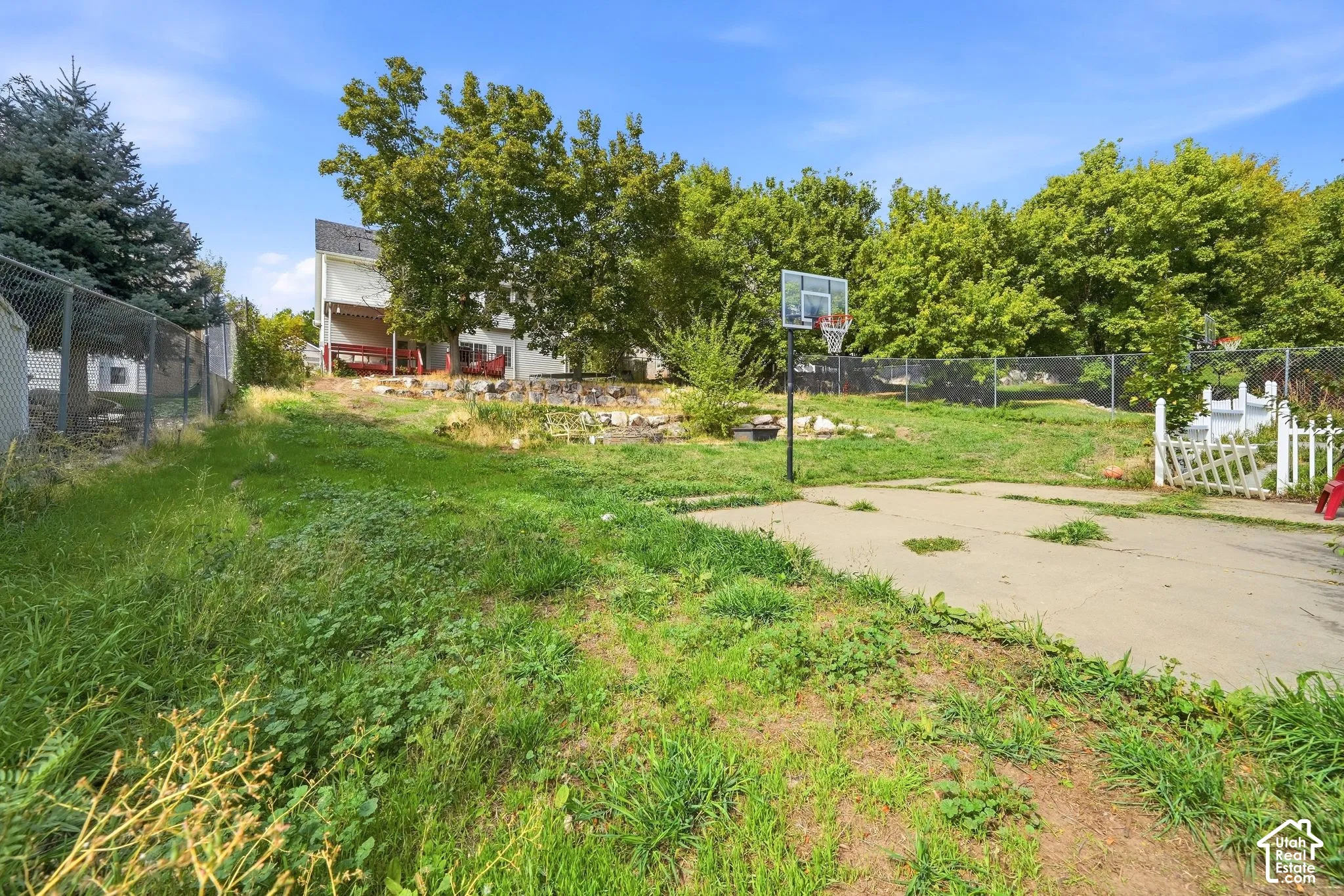 Paved court area with a basketball hoop, surrounded by established trees and a chain-link fence