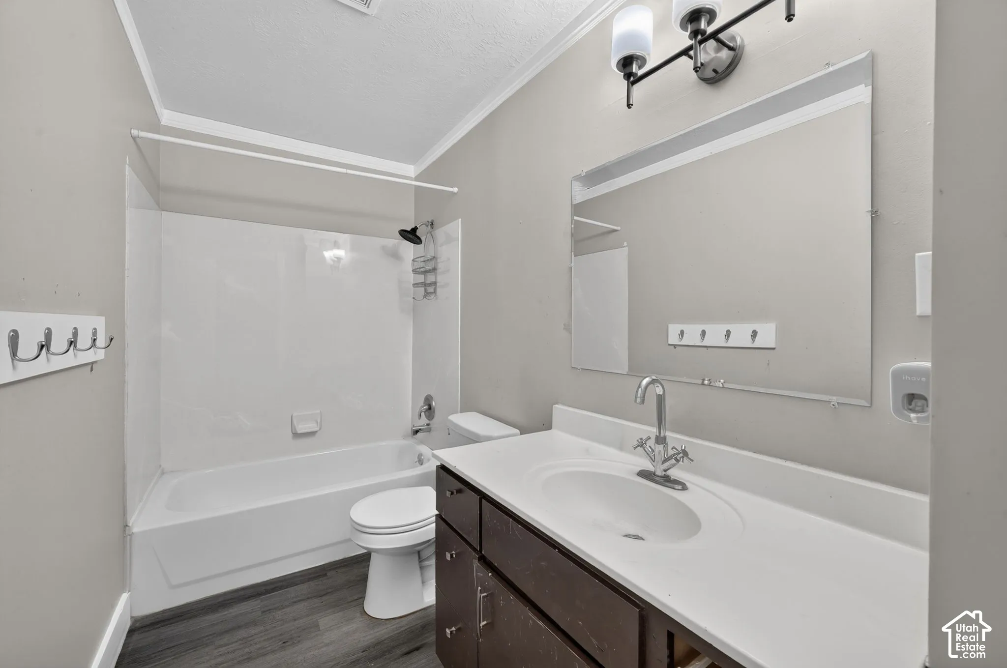 White bathtub with an integrated shower, dark wood-finish vanity with a white countertop and integrated sink, chrome-finish faucet, wall-mounted mirror, and wood-finish flooring