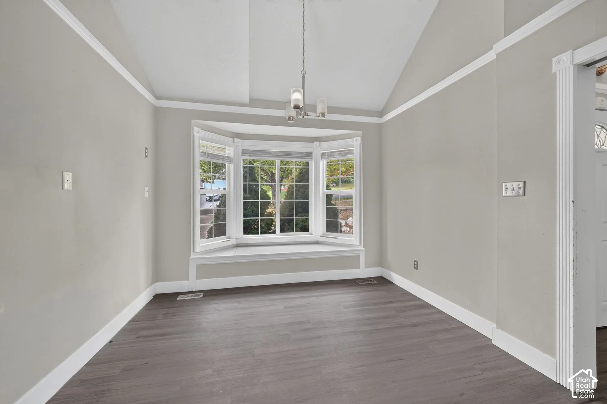 Vaulted ceiling with crown molding, bay window with integrated sill, wood-finish flooring, and a contemporary chandelier