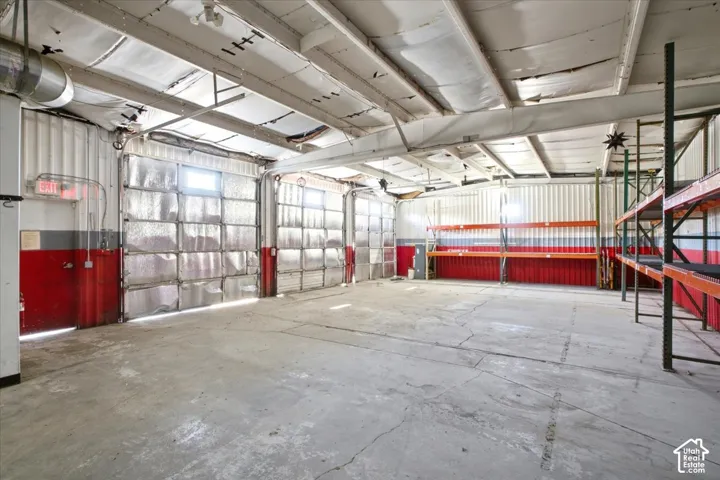 Concrete flooring, multiple roll-up doors with windows, exposed ceiling beams, built-in shelving, and wall-mounted exhaust fan
