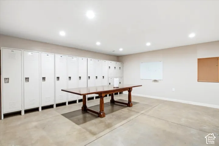 Recessed lighting illuminates the space featuring concrete flooring, a long bank of white lockers, a wood-finish central table, and a wall-mounted whiteboard