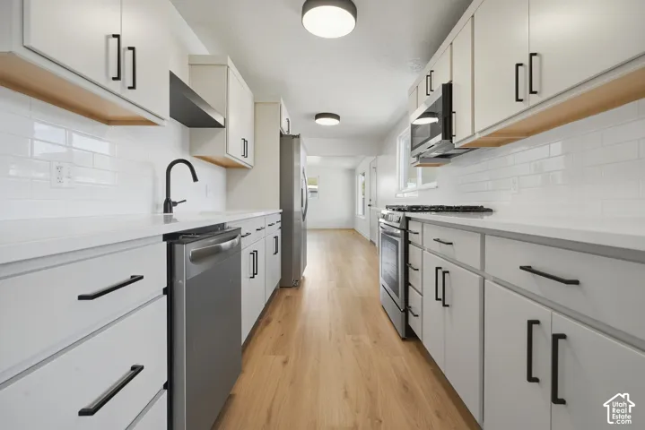 Galley kitchen featuring wood-finish flooring, white cabinetry with black hardware, white subway tile backsplash, white countertops, and stainless steel appliances