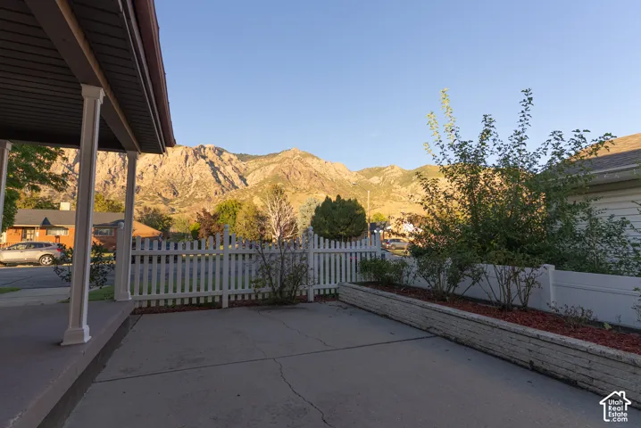 Paved driveway with a partial covered porch, white picket fencing, and a raised planter bed, complemented by a mountain backdrop