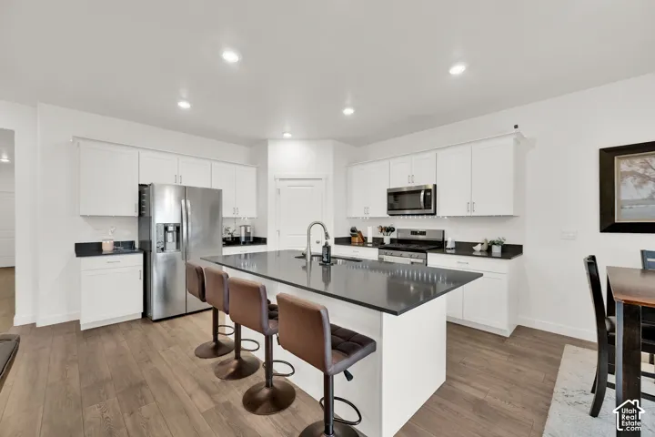 Kitchen featuring white shaker cabinetry, stainless steel appliances, dark countertops, a center island with sink, and wood-finish flooring