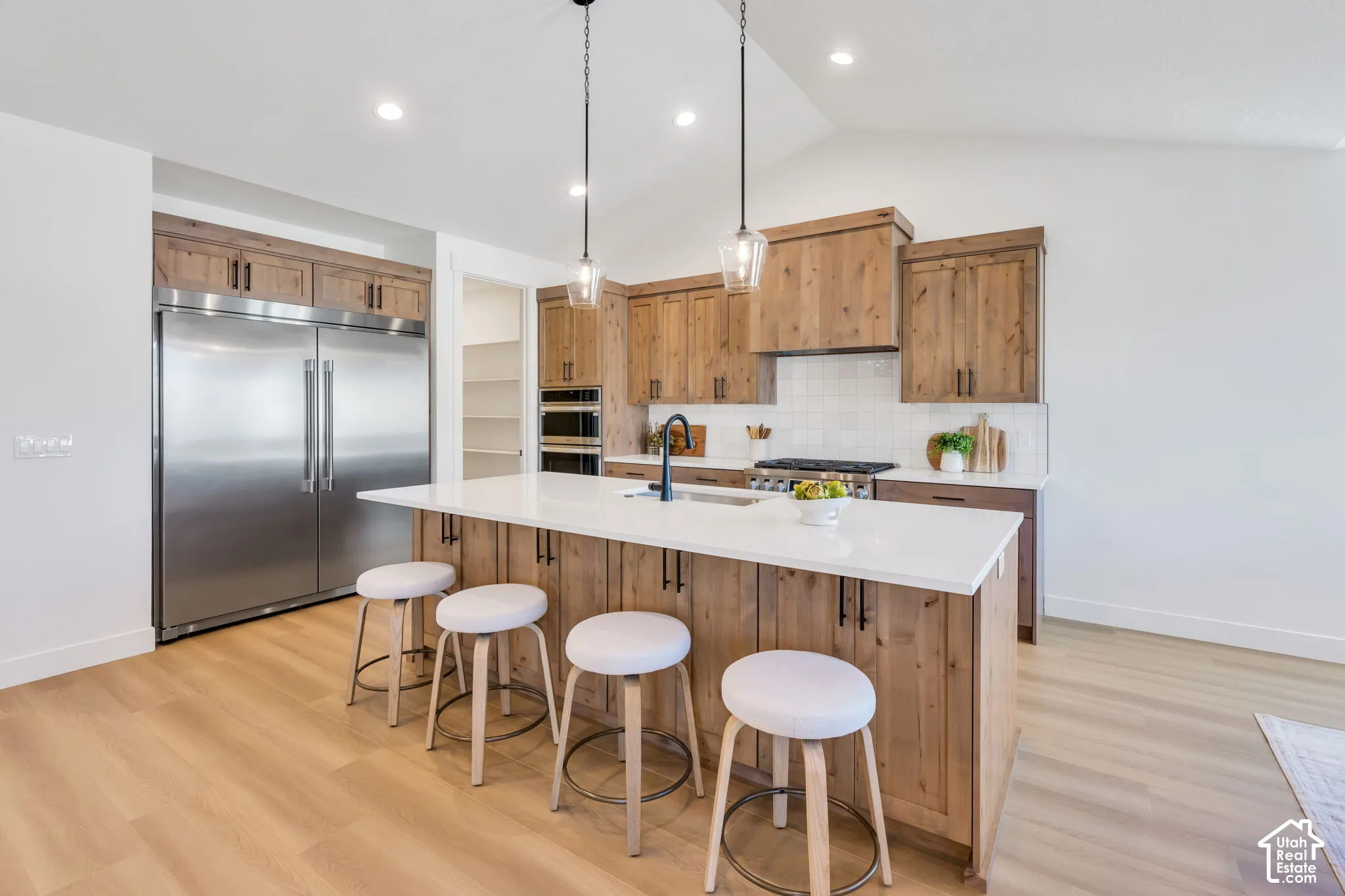 Contemporary kitchen featuring custom wood cabinetry, white quartz countertops, stainless steel appliances including a built-in refrigerator and dual wall ovens, a gas cooktop with hood, and a large waterfall-edge island with seating for four