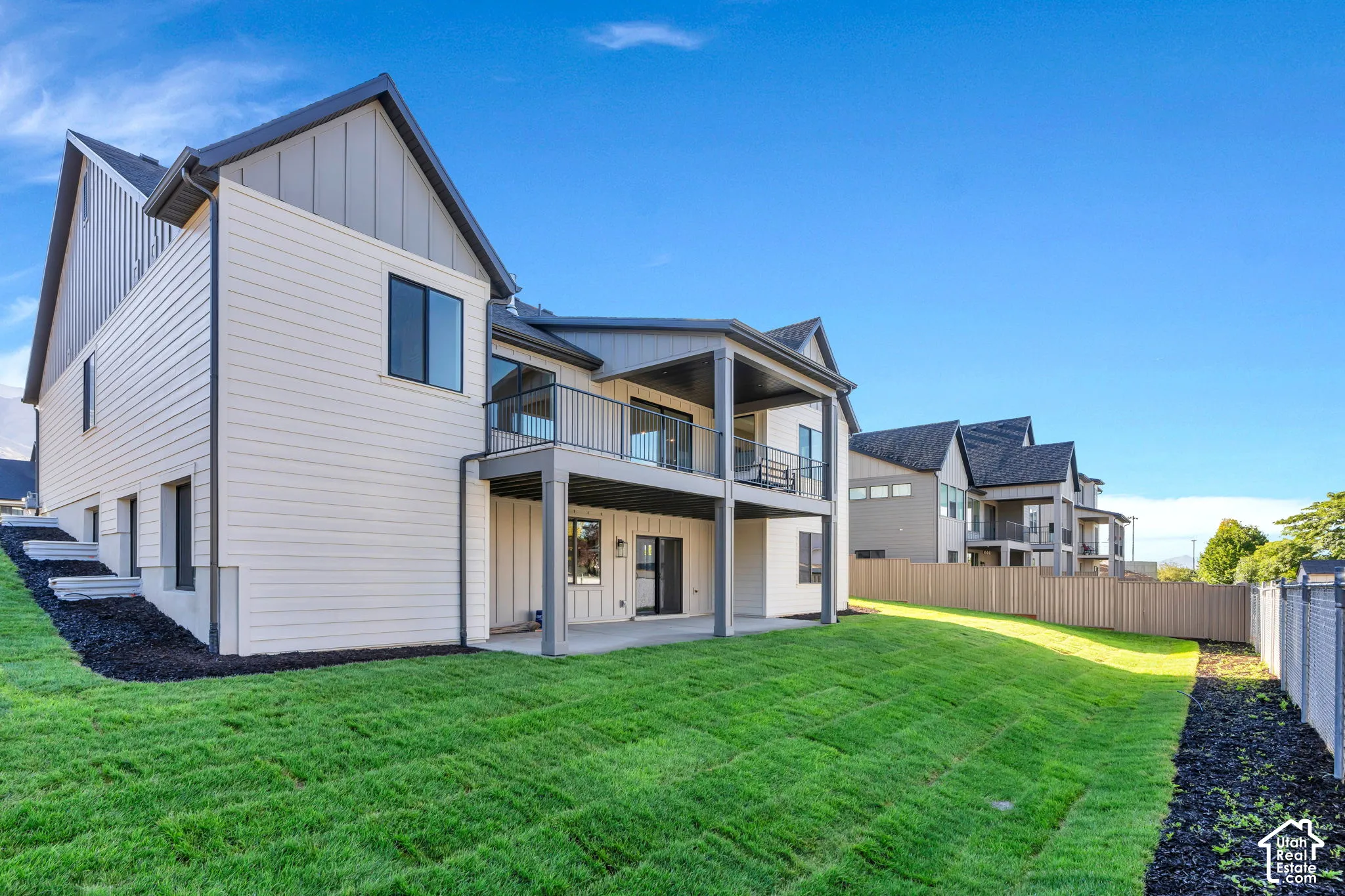 Multi-level exterior featuring white horizontal siding, vertical dark paneling, a covered balcony with dark metal railings, a ground-level covered patio, and a sloped grass lawn