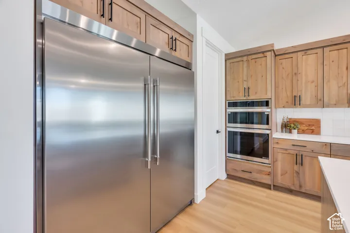 Stainless steel refrigerator, integrated double wall ovens, wood-finish cabinetry with black hardware, white tile backsplash, and light wood-finish flooring