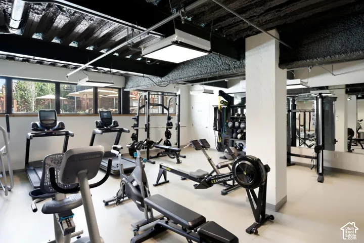 Fitness center with expansive windows, exposed ceiling beams, built-in lighting, wall-mounted mirrors, and durable flooring