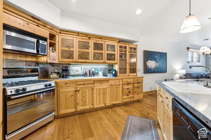 Wood-finish flooring, stainless steel appliances, stone tile backsplash, wood cabinetry with textured glass inserts, and a built-in dishwasher